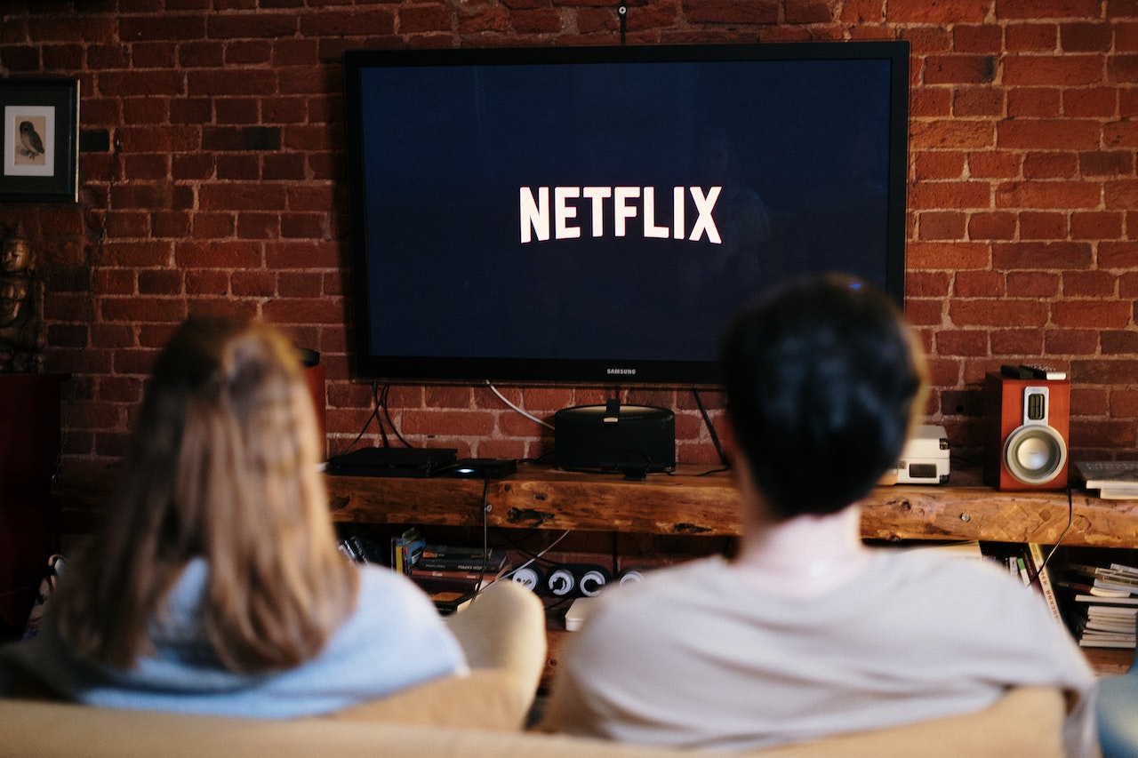 På bilden kan man se två personer som sitter på en soffa och tittar på en TV där Netflix-logotypen visas. Bakom TV:n finns en tegelvägg. Det finns några dekorativa objekt, högtalare och böcker runt TV:n