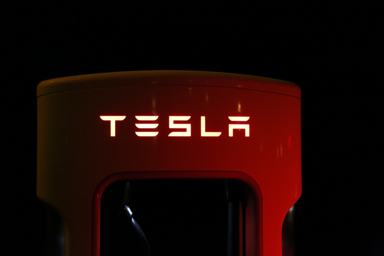 Bilden visar en del av en Tesla-laddstation, som troligtvis används för att ladda Tesla-elbilar. Företagets välkända logotyp "TESLA" är framträdande och belyst i en iögonfallande röd färg. Denna röda belysning kontrasterar starkt mot den mörka bakgrunden, vilket ger en kraftfull visuell effekt. Designen och belysningen antyder Tesla's moderna och innovativa varumärke. Det är tydligt att detaljerna och kvaliteten på stationen är hög, vilket reflekterar företagets engagemang i premiumprodukter